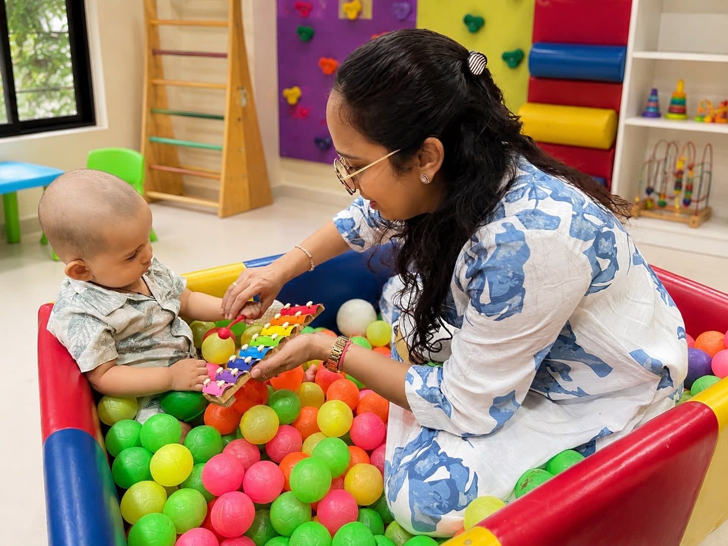 Baby playing a colorful xylophone in the ball pit with Dr. Sharuja during a sensory play session