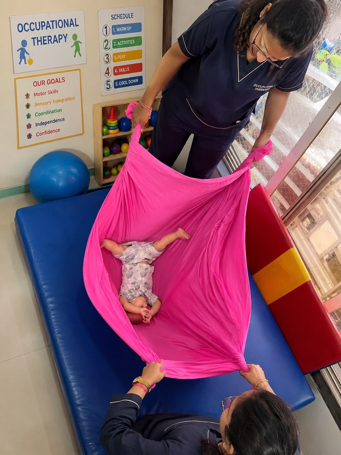 Infant relaxing in a soft pink blanket swing held by two therapists for vestibular input