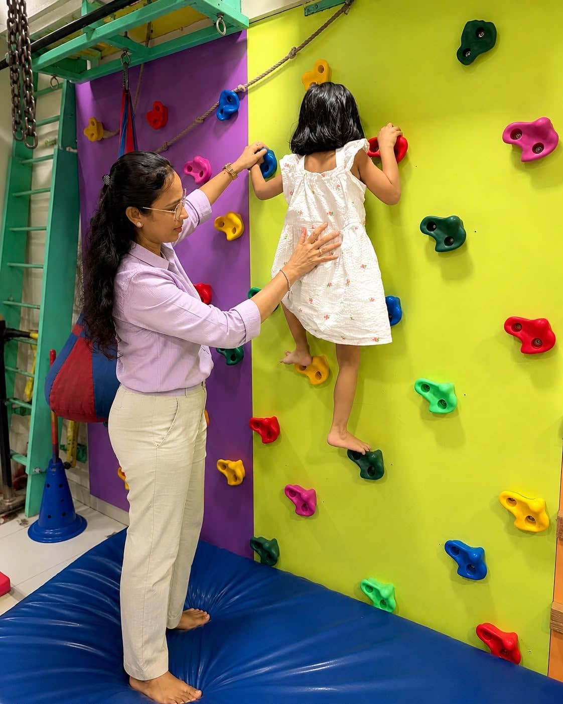 Young girl climbing a colorful climbing wall with close therapist support for motor planning and strength