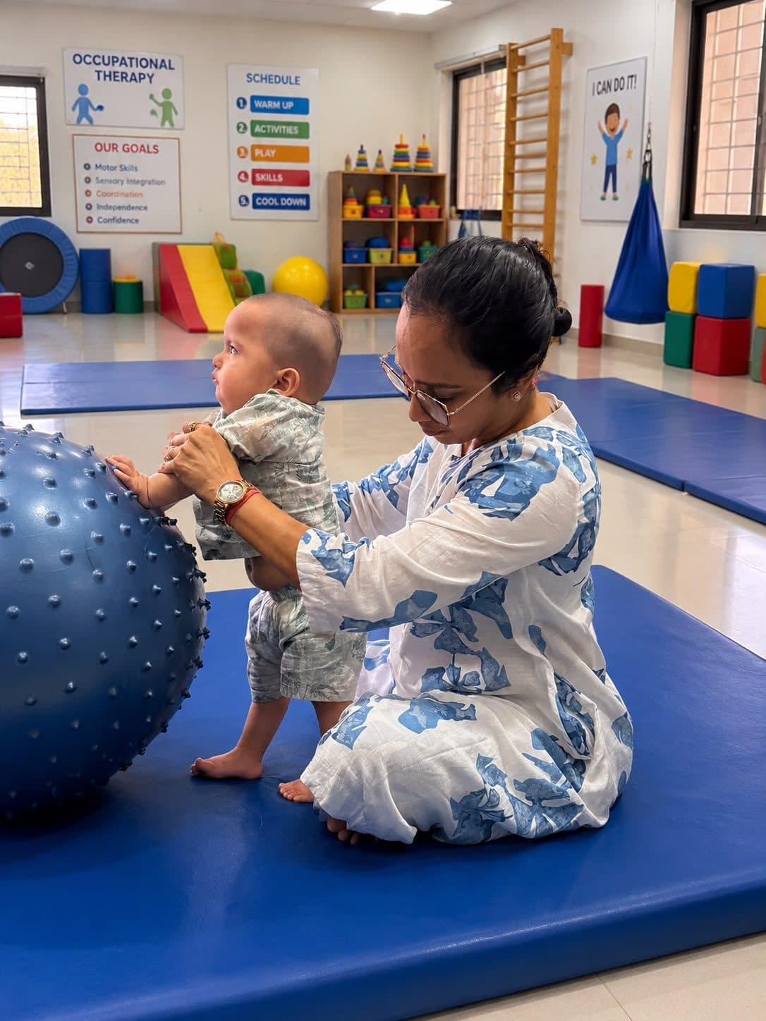 Infant practicing supported standing at a textured therapy ball in the spacious pediatric gym