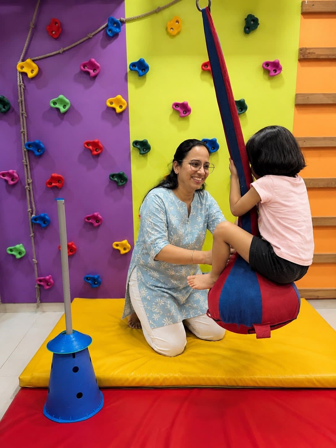 Girl seated on a therapy swing beside the climbing wall with a smiling therapist providing support